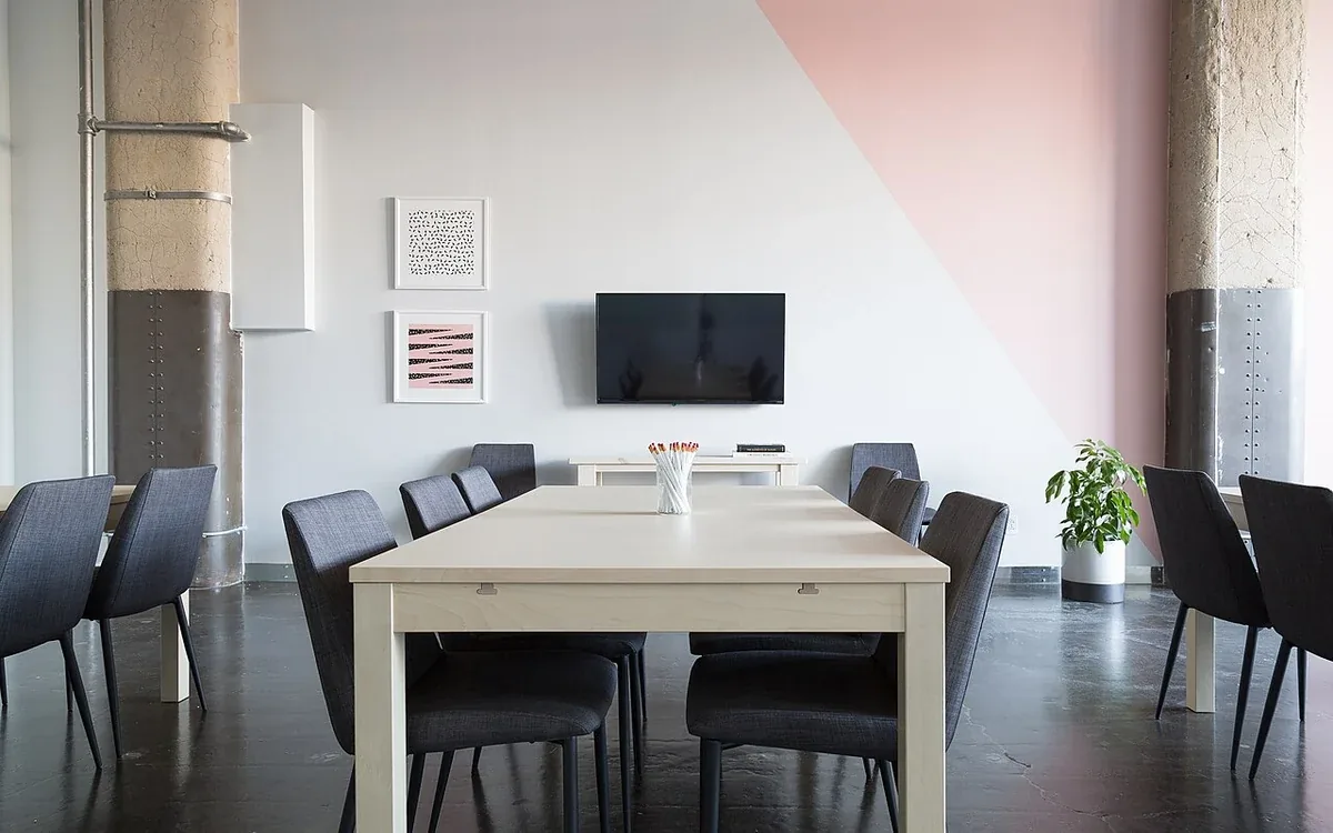 A light wooden table with dark chairs stands in a pale room beside windows, wall artwork and a plant