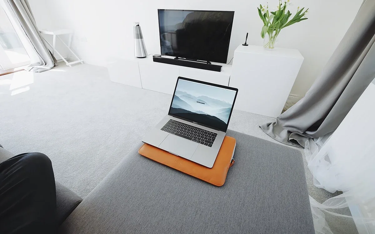 An open laptop rests on an orange mat on a gray upholstered seat, with a television and pale cabinet beyond