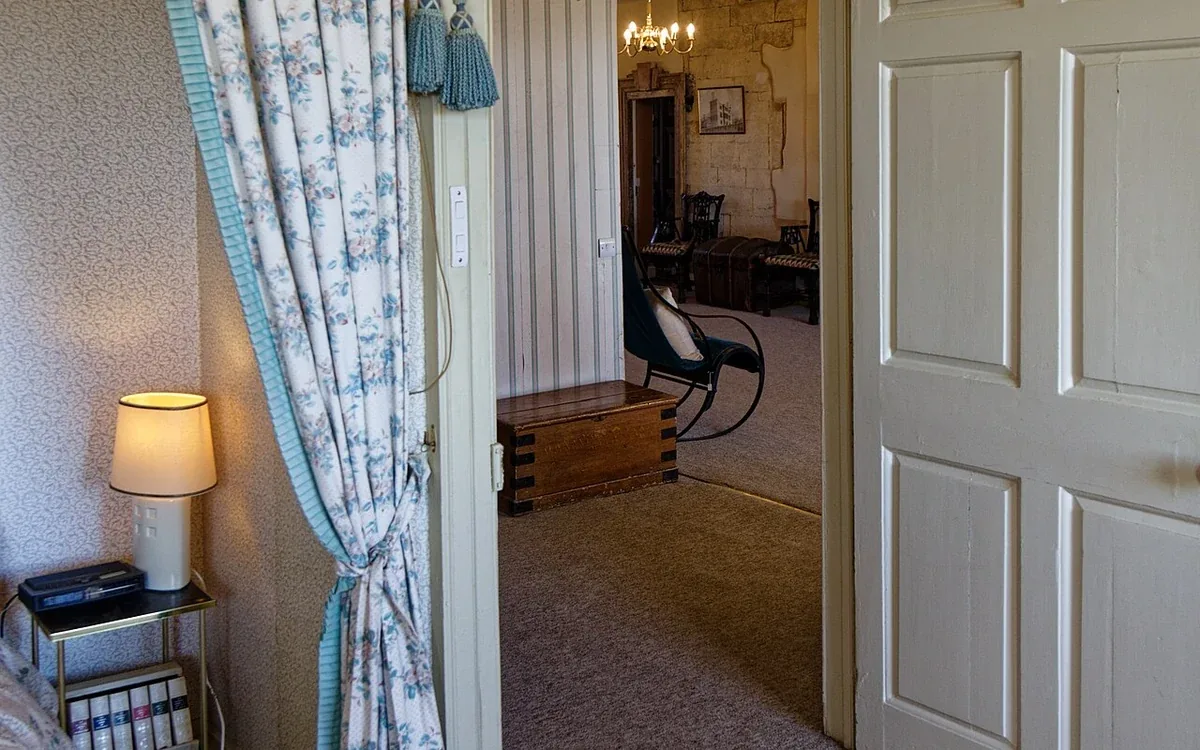 An open white door reveals part of a furnished bedroom beyond patterned curtains and a carpeted floor