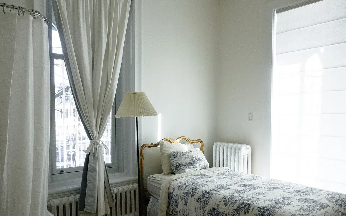 A bed with a pale patterned cover stands beside a tall window, gray curtains and a floor lamp in a bright room