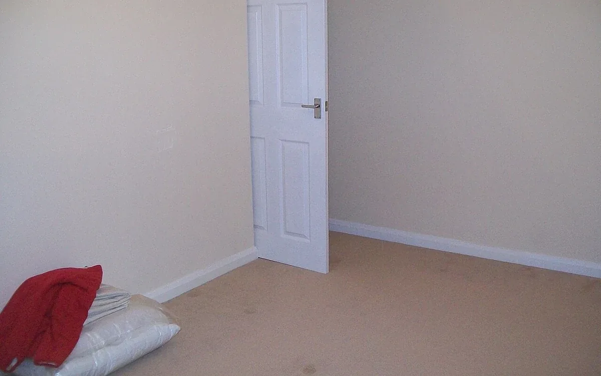 A pale-walled bedroom has a closed white door, a clear carpeted floor and a small pile of bedding at one side