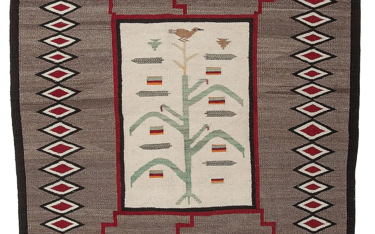 A red and cream woven rug features a central plant motif and angular patterns inside a dark border