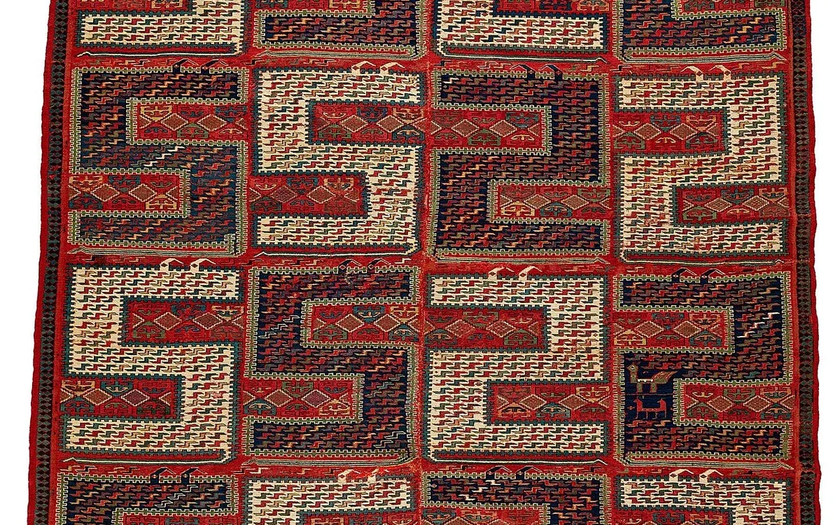 A red and dark flatweave rug with interlocking geometric shapes lies flat against a plain light background