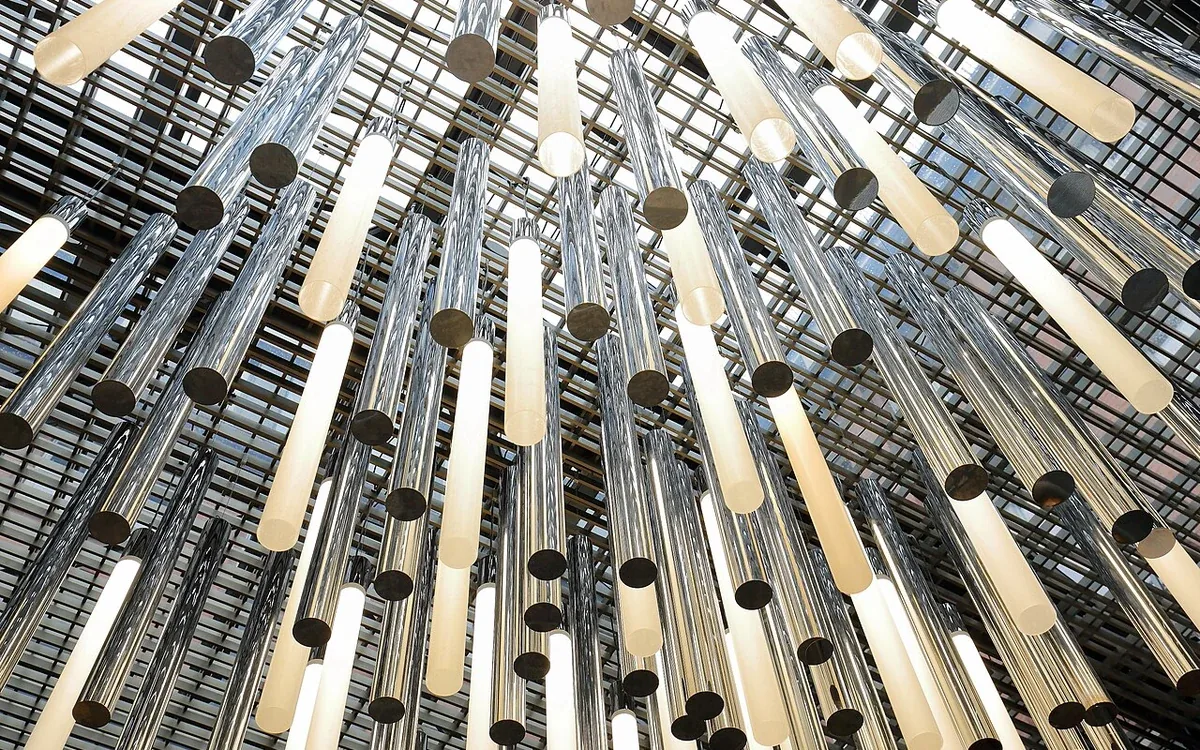 Long illuminated tubes and reflective metal cylinders hang beneath a metal ceiling grid, viewed from below