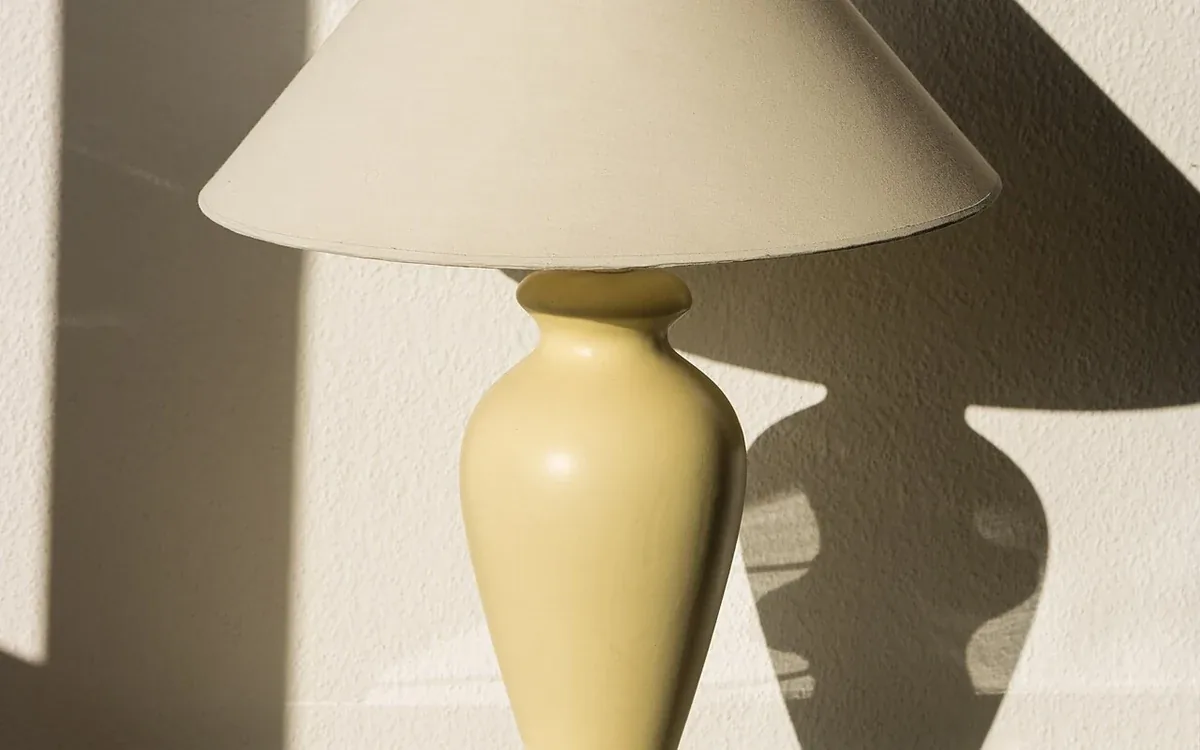 A pale ceramic table lamp with a wide fabric shade stands on a reflective surface in sunlight