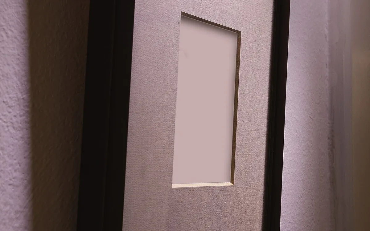 A dark rectangular picture frame with a light inner mount hangs on a pale wall and casts a soft shadow