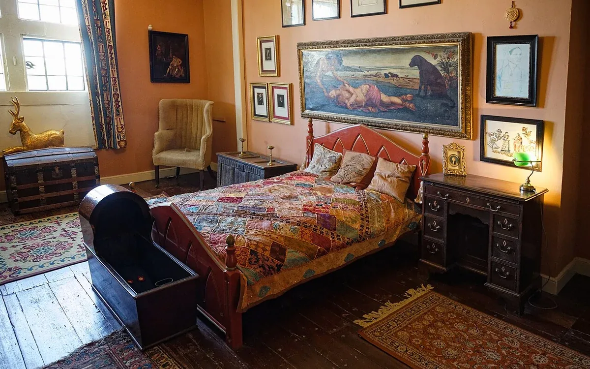 A bed with a red patterned cover sits between wooden bedside furniture beneath several framed pictures