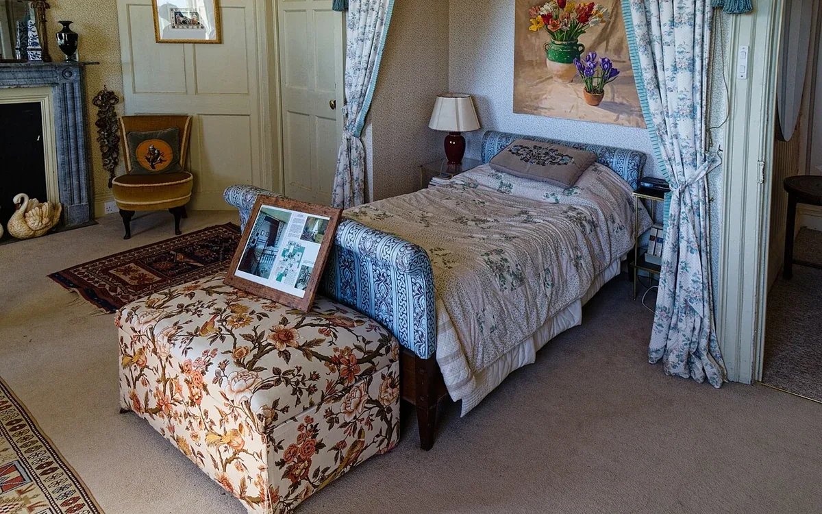 A bed with patterned covers stands near an upholstered stool, wooden chairs and framed pictures in a furnished bedroom