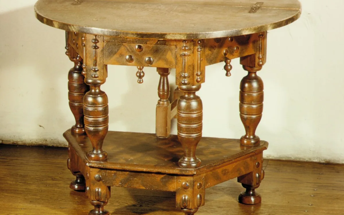 Ornate brown wooden table with a rounded hinged top, turned legs and a lower shelf beside a pale wall.