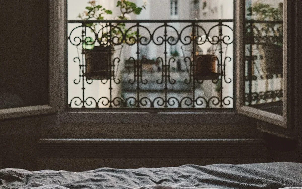 An open window looks toward a balcony rail and nearby buildings above the edge of a bed with rumpled covers