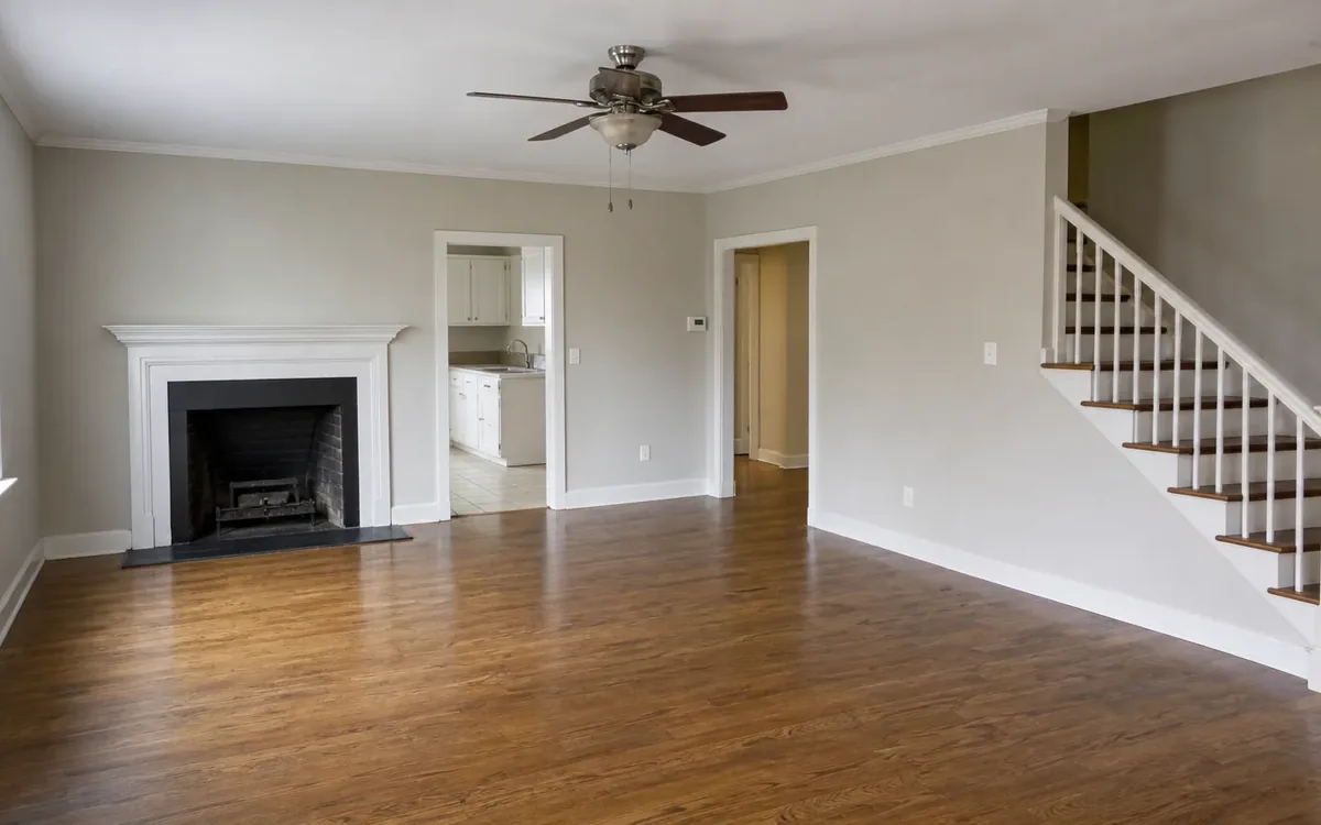 Empty living room with hardwood floors, a fireplace, ceiling fan, kitchen doorway, and staircase after move-out.