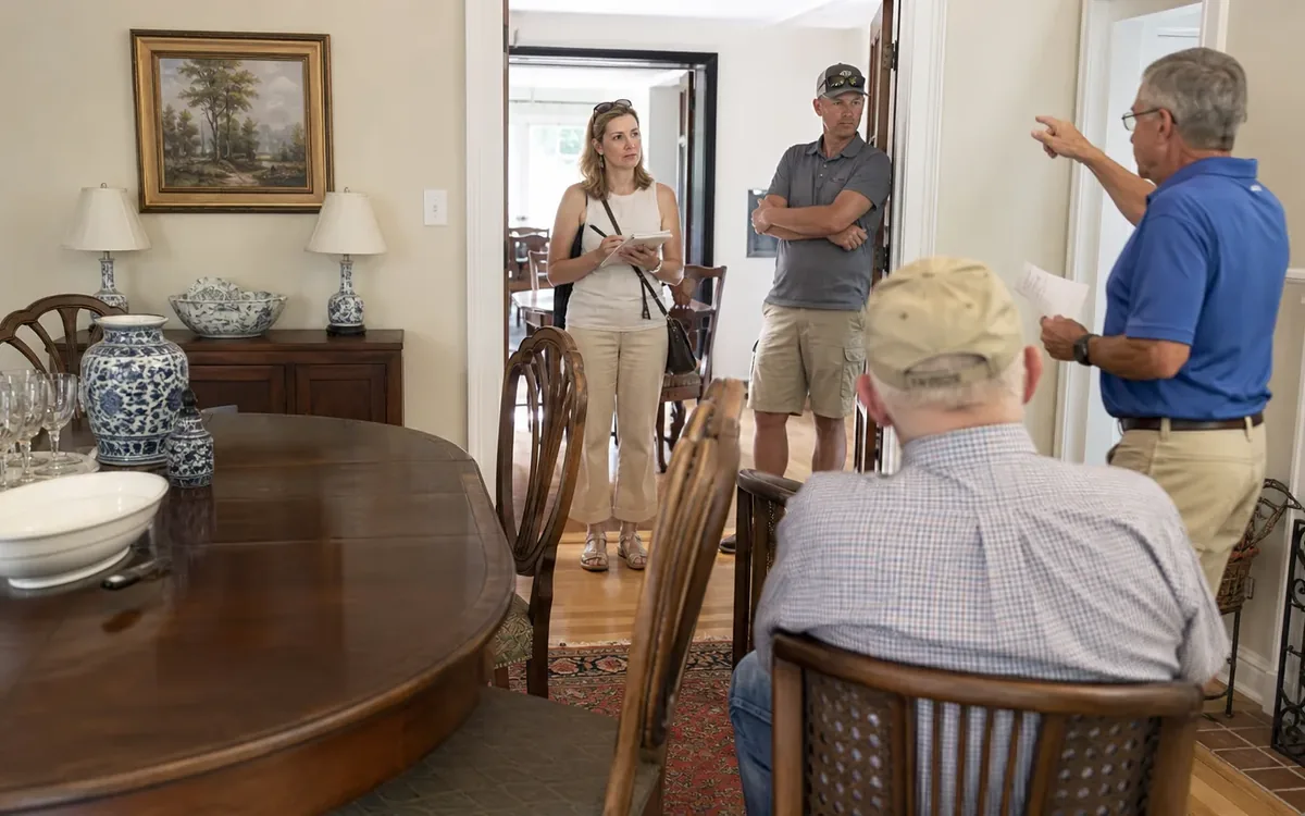 A man gestures to three people in a dining room furnished with a wooden table, chairs, glassware, and blue-and-white ceramics.