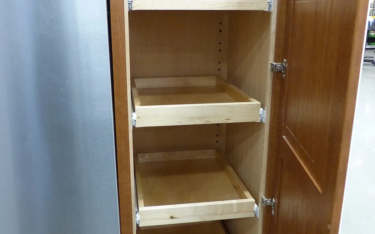An open tall wooden cupboard contains several empty shelves and a lower pull-out frame beside a white wall