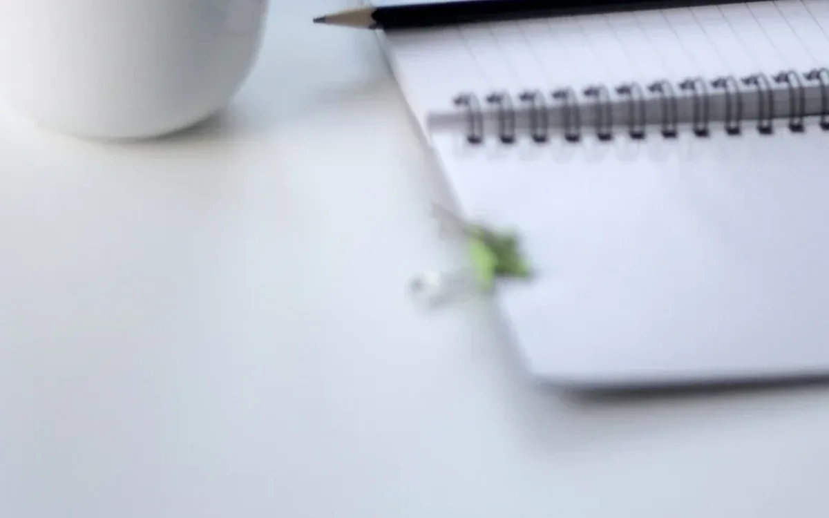 A spiral notebook with blank ruled pages lies beside a dark pen and part of a white mug on a pale desk