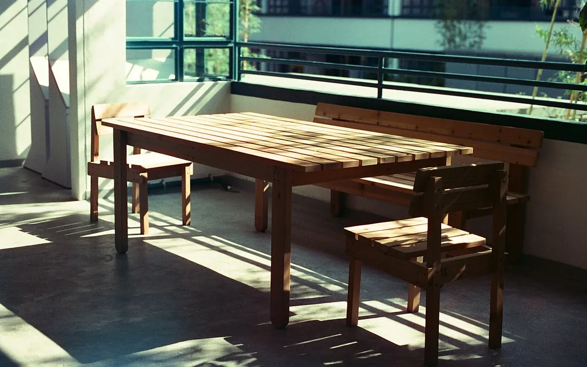 A plain wooden table and matching benches stand beside large windows in a sunlit room