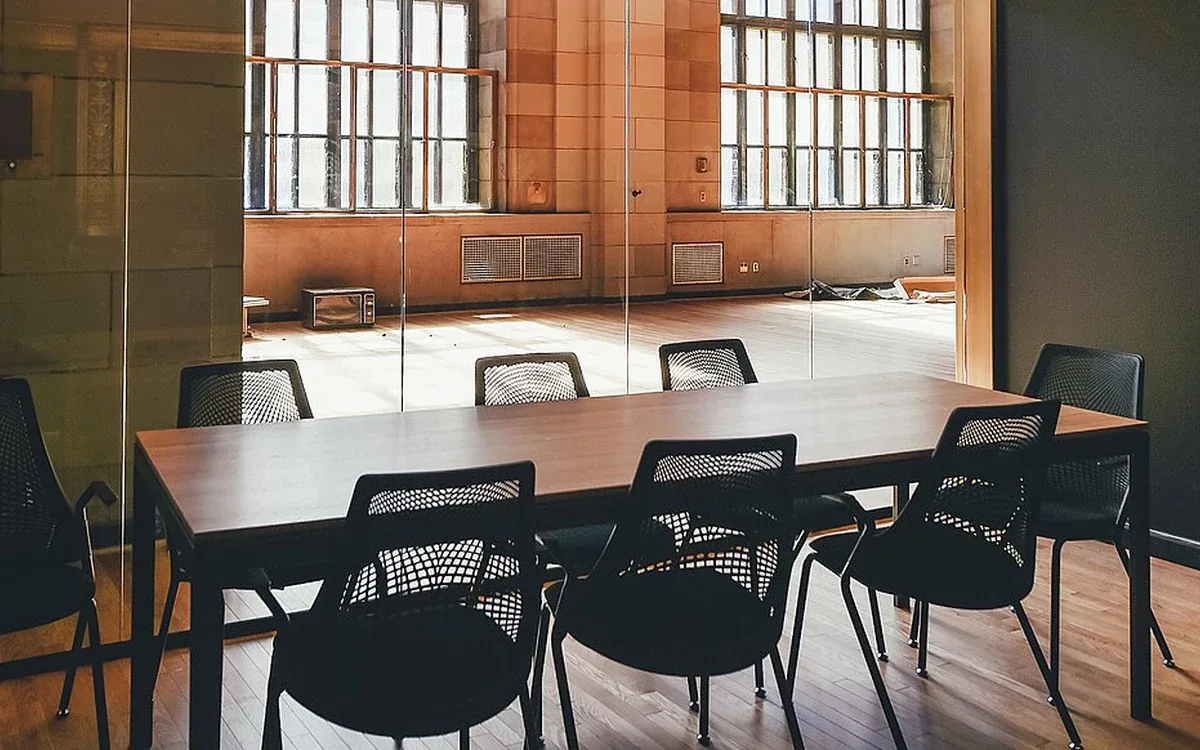 A rectangular table and dark chairs stand on a wooden floor beside large windows and exposed brickwork