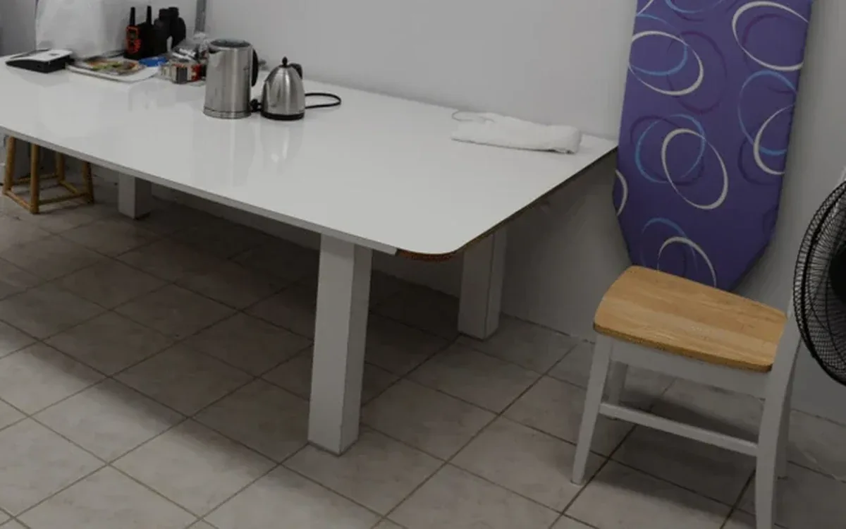 A clear white table and light chairs stand on a tiled floor beside laundry equipment and a pale wall