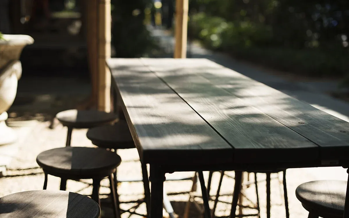 A broad wooden table and several stools stand beside plants and open space