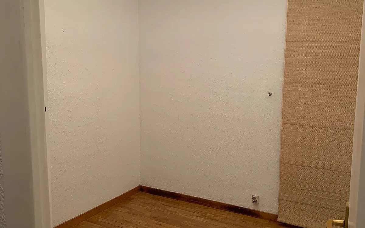 An empty white room has wooden flooring, a built-in wooden cupboard and bare light bulbs hanging from the ceiling