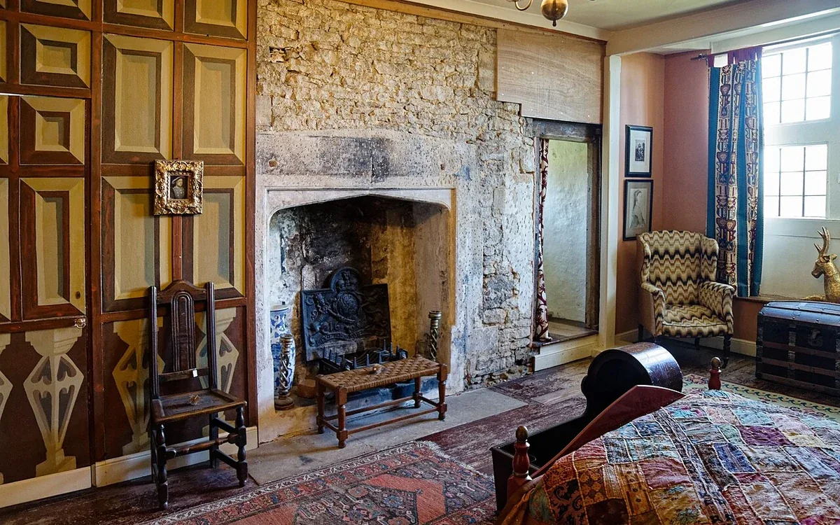 A broad stone fireplace stands beside chairs, framed artwork and patterned bedding in a furnished room