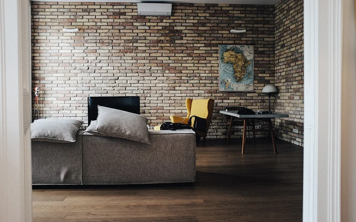 A gray sofa with cushions stands beneath a brick wall, beside a dining table in an open room with wooden flooring