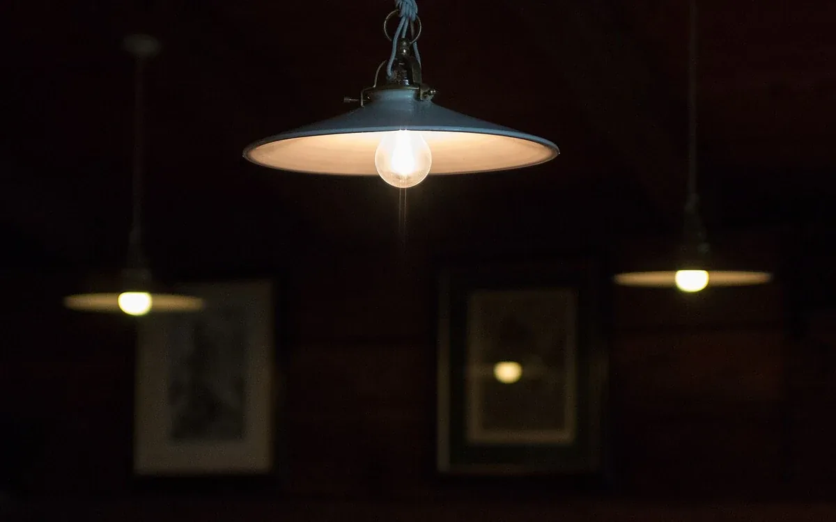 A wide metal pendant light hangs above a dim interior, with framed pictures and other lights blurred behind it
